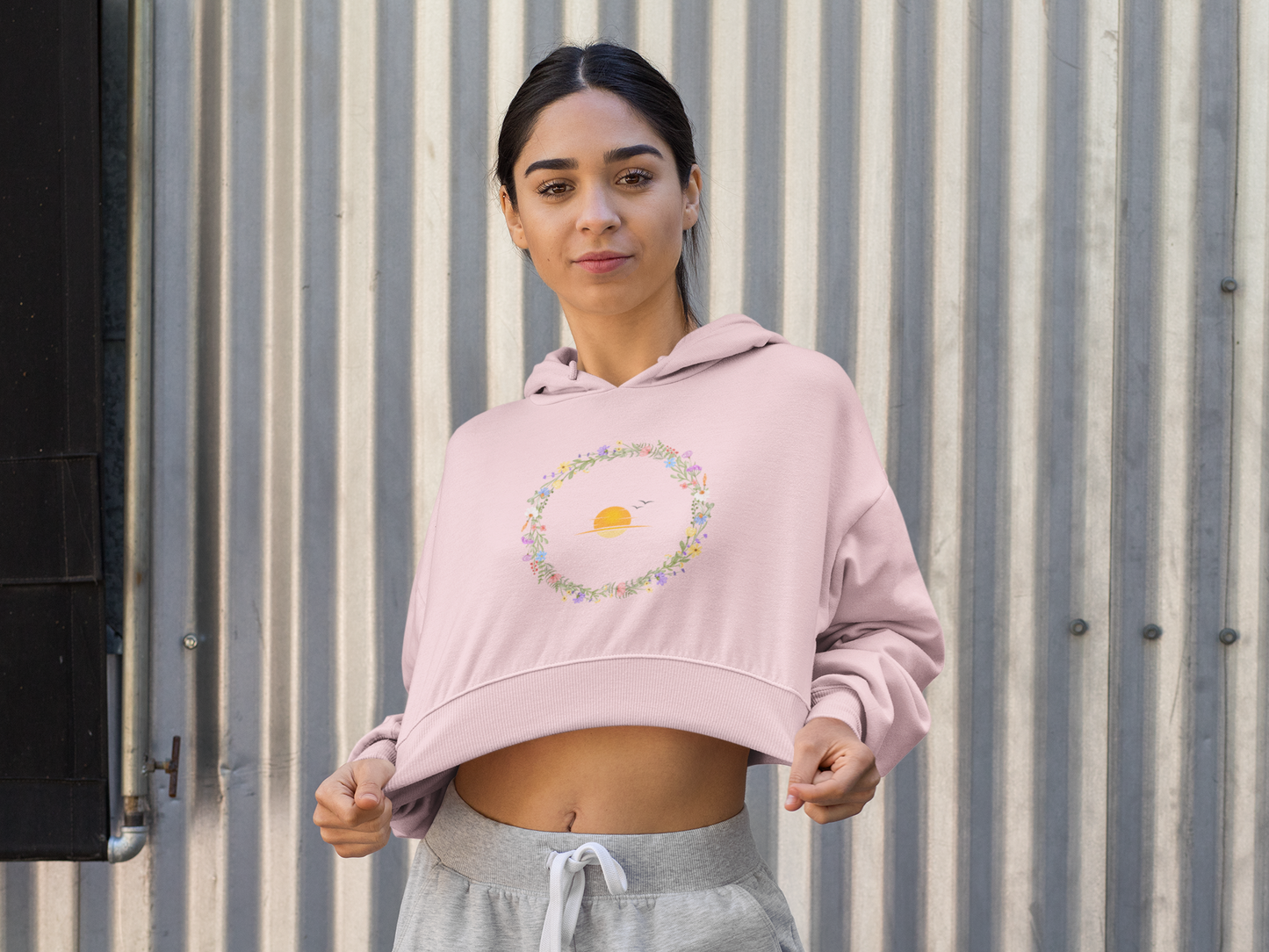 Light Baby Pink Woman wearing a pink hoodie with a floral design and sun graphic, standing against a corrugated metal wall.