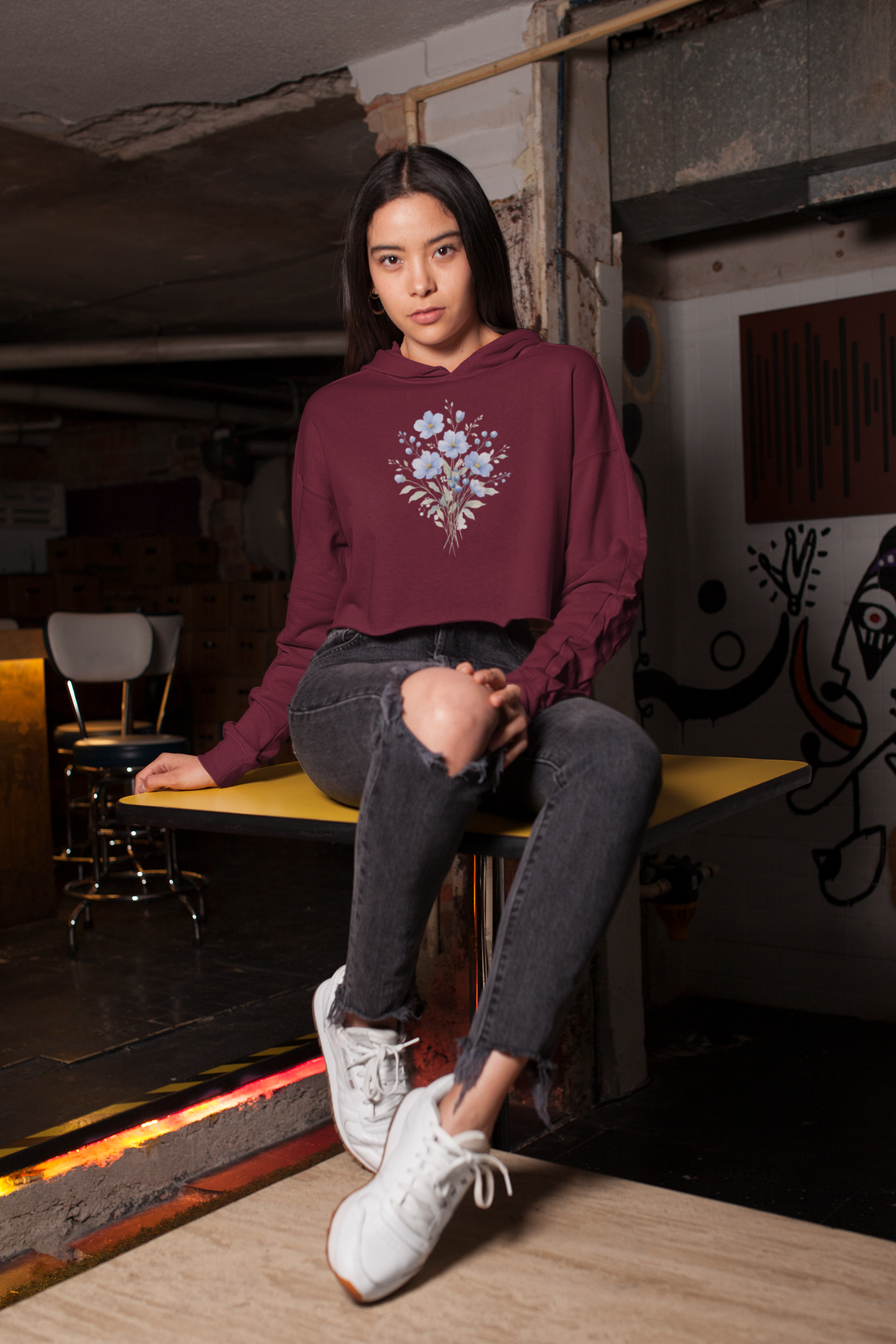 Woman wearing a maroon hoodie with floral design sitting on a bench in an urban setting.