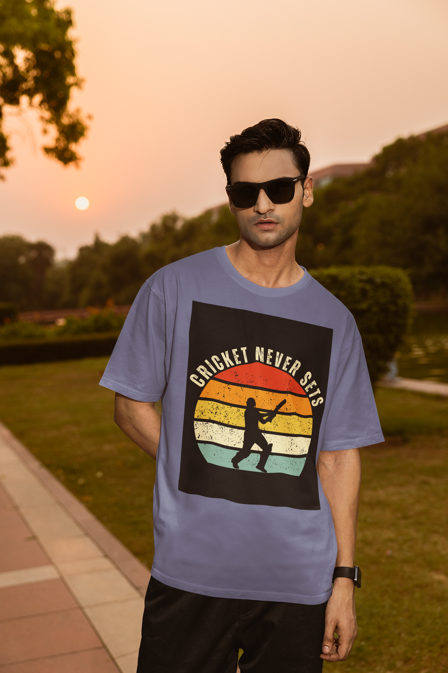Man wearing a steel grey t-shirt with 'Cricket Never Sleeps' design outdoors during sunset.