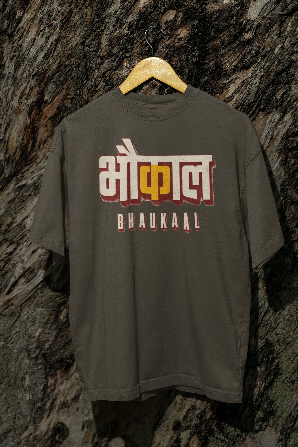 Olive Green Bhaukaal oversized t-shirt on a hanger against a textured bark wall, showing the full 3D Hindi typography front print