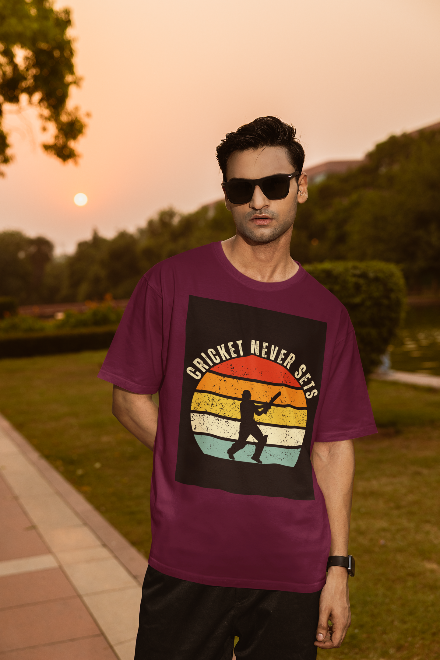 Man wearing a burgundy t-shirt with 'Cricket Never Sleeps' design outdoors during sunset.