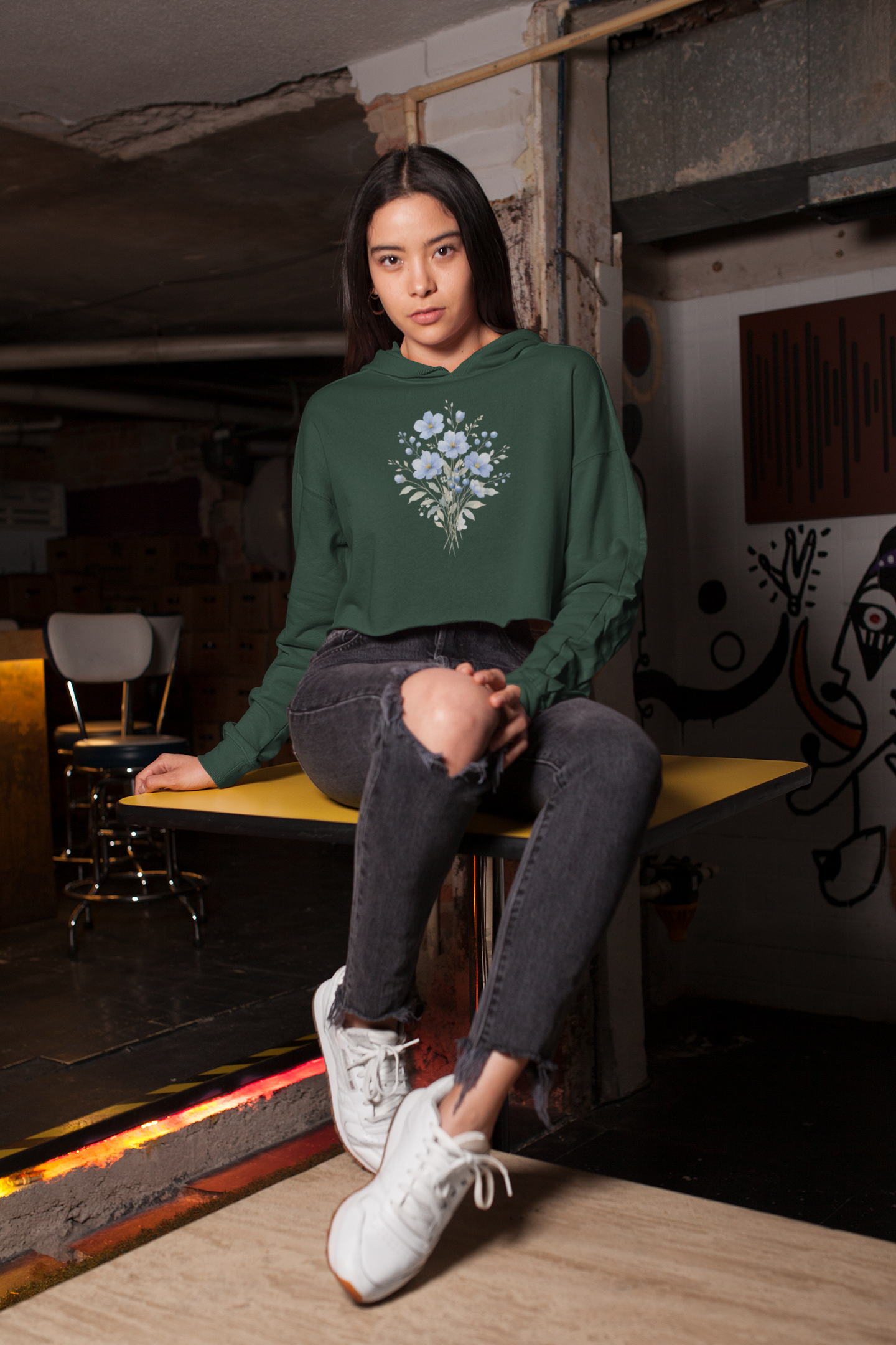 Woman wearing a olive green hoodie with floral design sitting on a bench in an urban setting.