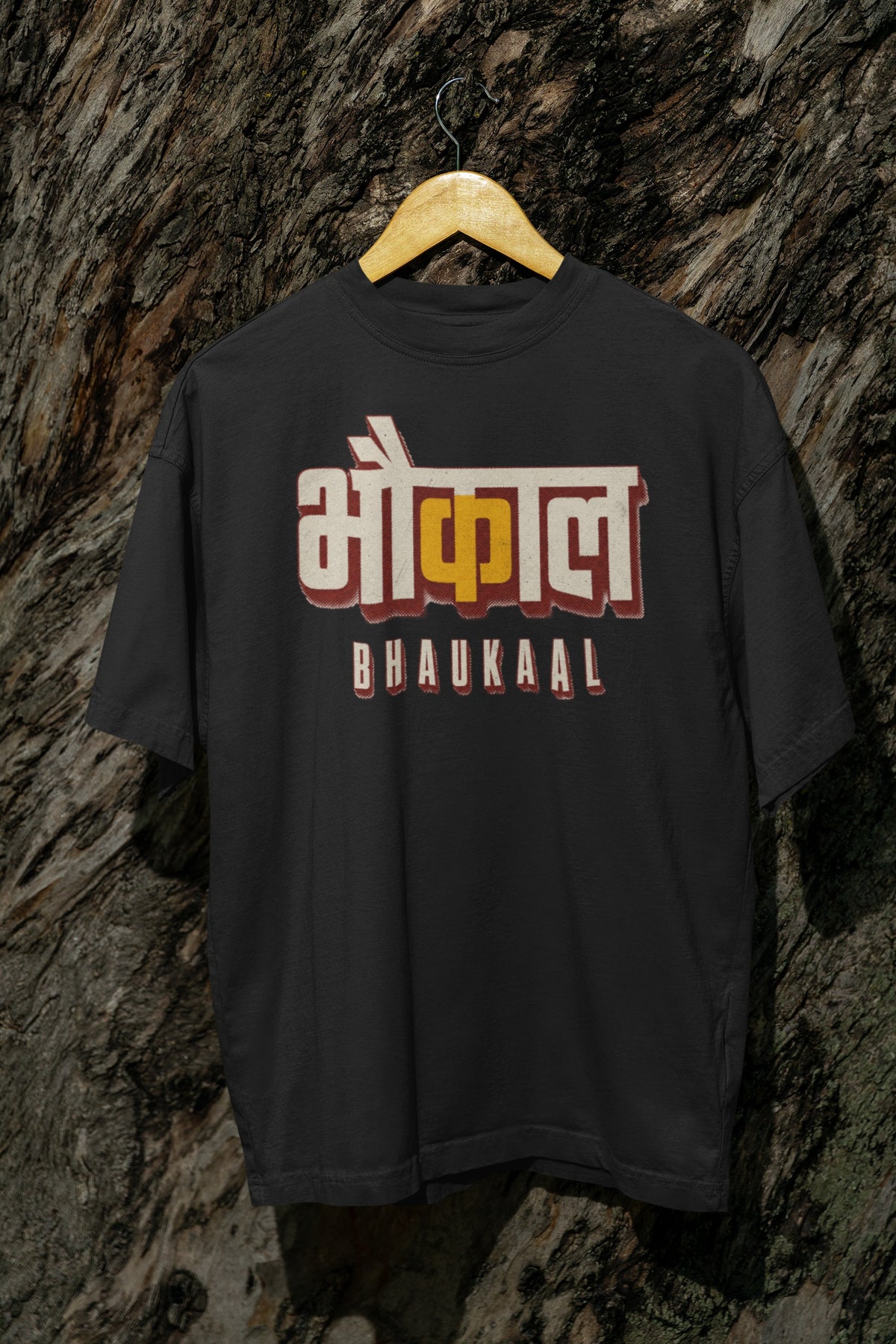 Black Bhaukaal oversized t-shirt on a hanger against a textured bark wall, showing the full 3D Hindi typography front print