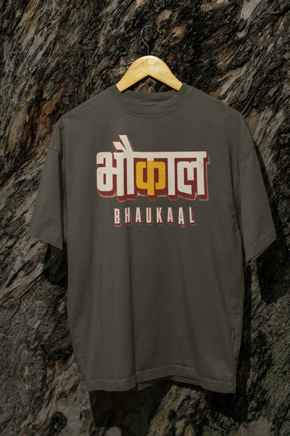 Olive Green Bhaukaal oversized t-shirt on a hanger against a textured bark wall, showing the full 3D Hindi typography front print