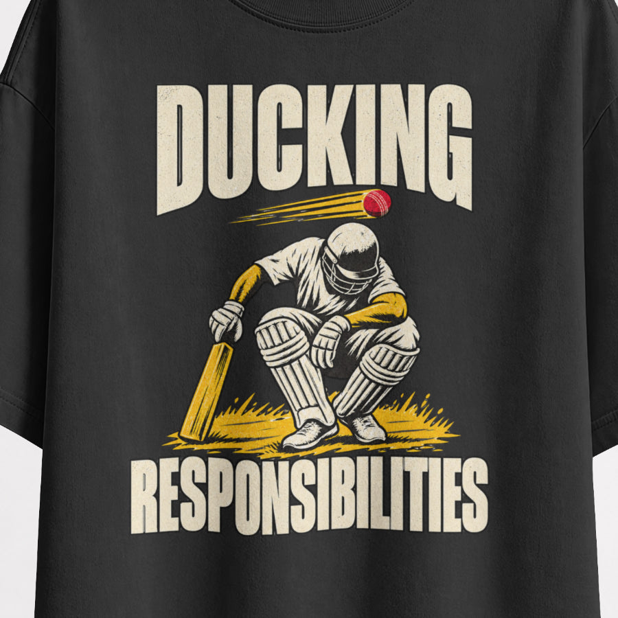 Black Ducking Responsibilities oversized T-shirt print close-up, cream DUCKING lettering over a helmeted cricket batter crouching under a red ball, mustard-yellow grass