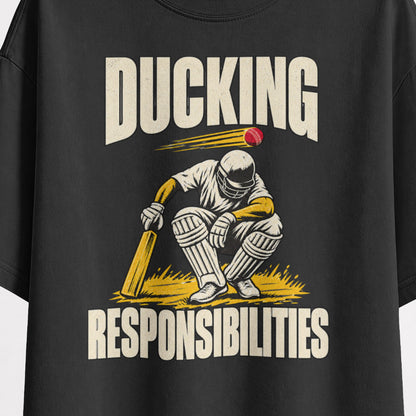 Black Ducking Responsibilities oversized T-shirt print close-up, cream DUCKING lettering over a helmeted cricket batter crouching under a red ball, mustard-yellow grass