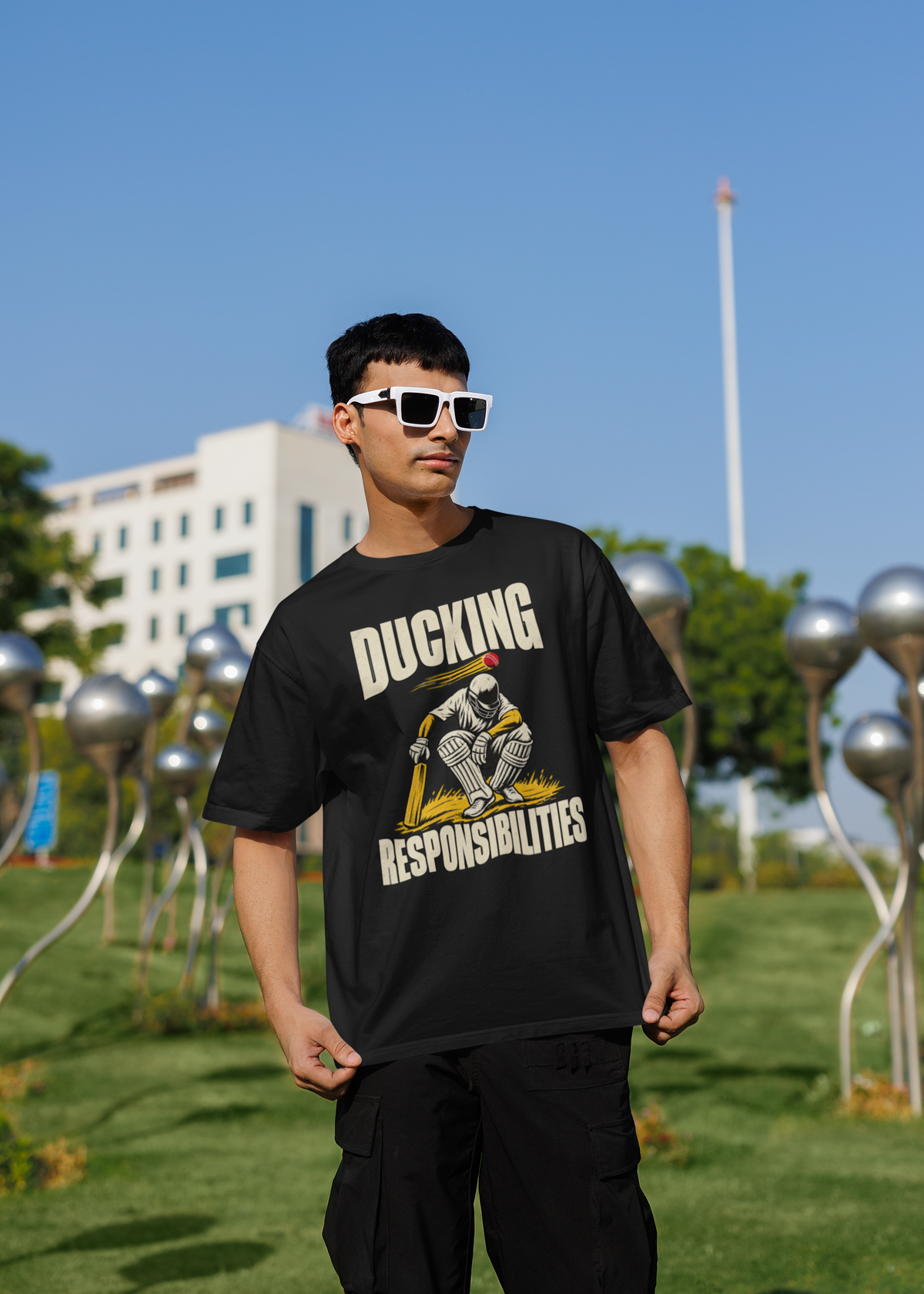 Man wearing the black Ducking Responsibilities oversized cricket T-shirt with black cargo trousers and white sunglasses, standing outdoors in a park