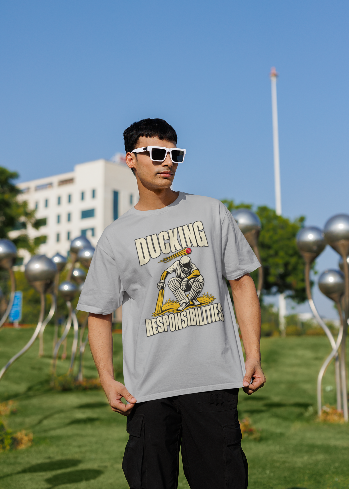 Man wearing the melange grey Ducking Responsibilities oversized cricket T-shirt with black cargo trousers, standing outdoors in a park