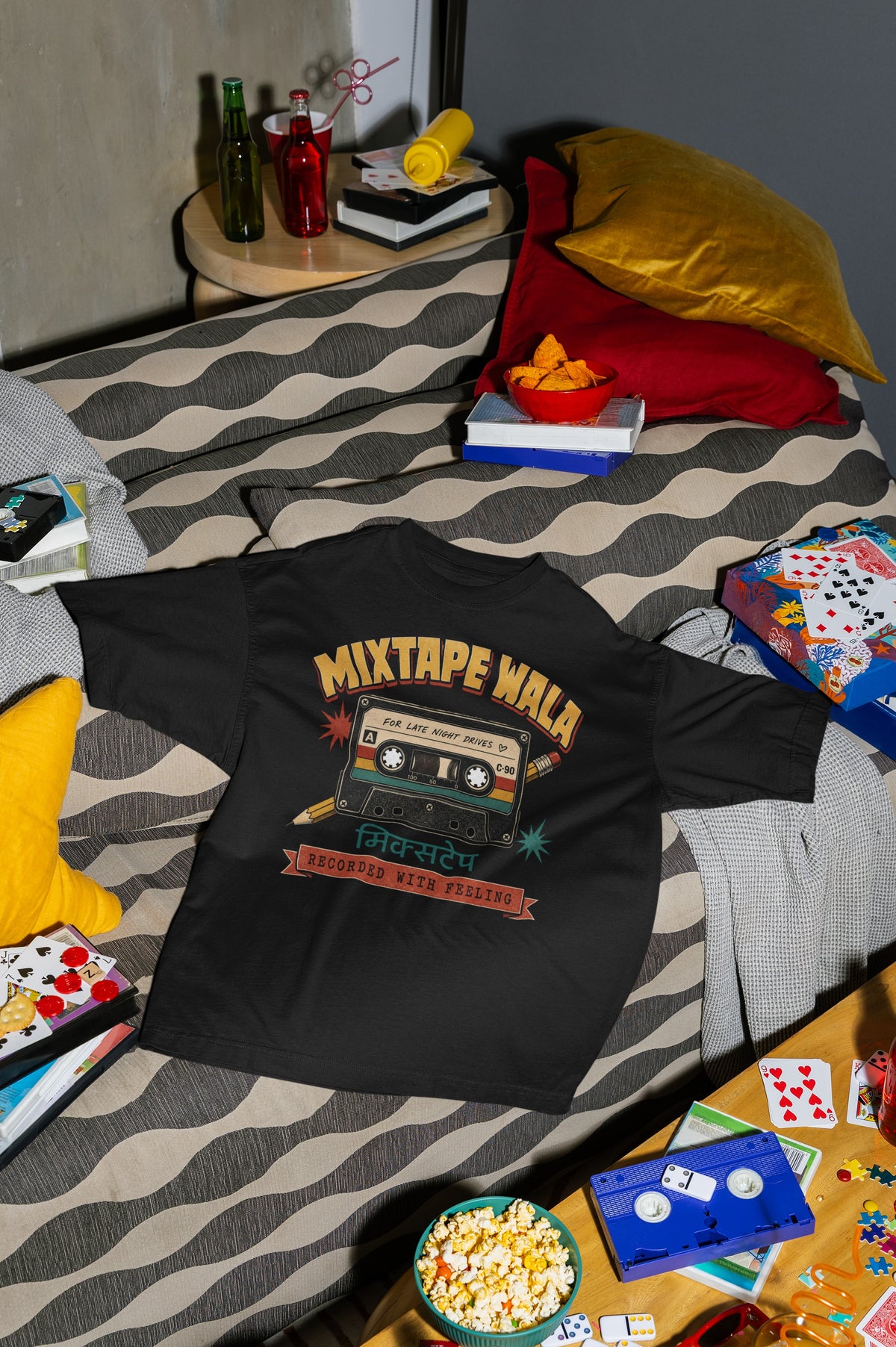 Black Mixtape Wala oversized t-shirt laid flat on a bed with cushions, cassette and popcorn