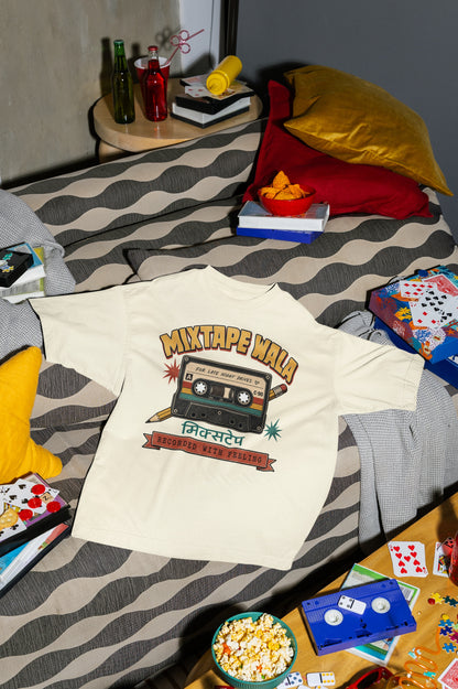 Off white Mixtape Wala oversized t-shirt laid flat on a bed with cushions, cassette and popcorn