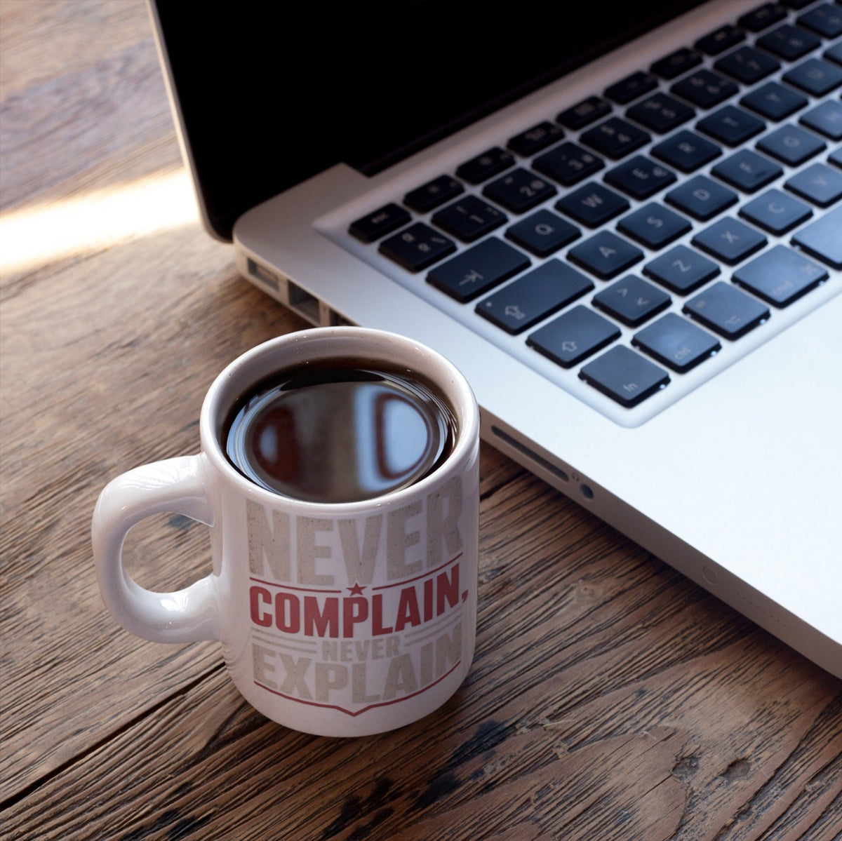 Never Complain Never Explain 11oz ceramic coffee mug styled on a work desk with a laptop